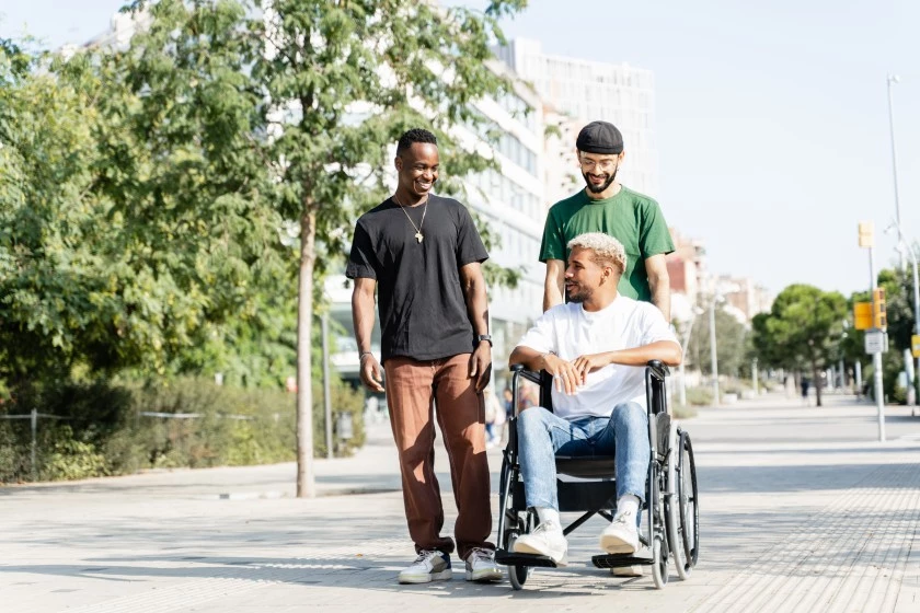 Three male friends, one man in wheelchair, walking outside and talking and smiling together